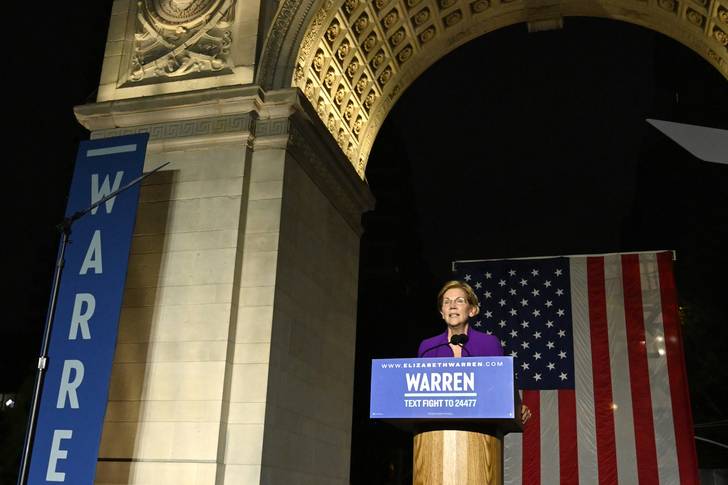 Elizabeth Warren standing on a stage in front of the Washington Square Park arch.